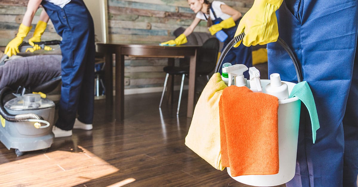 Integrity Cleaning Services team in blue uniforms and yellow gloves cleaning a living room, holding a bucket of cleaning supplies